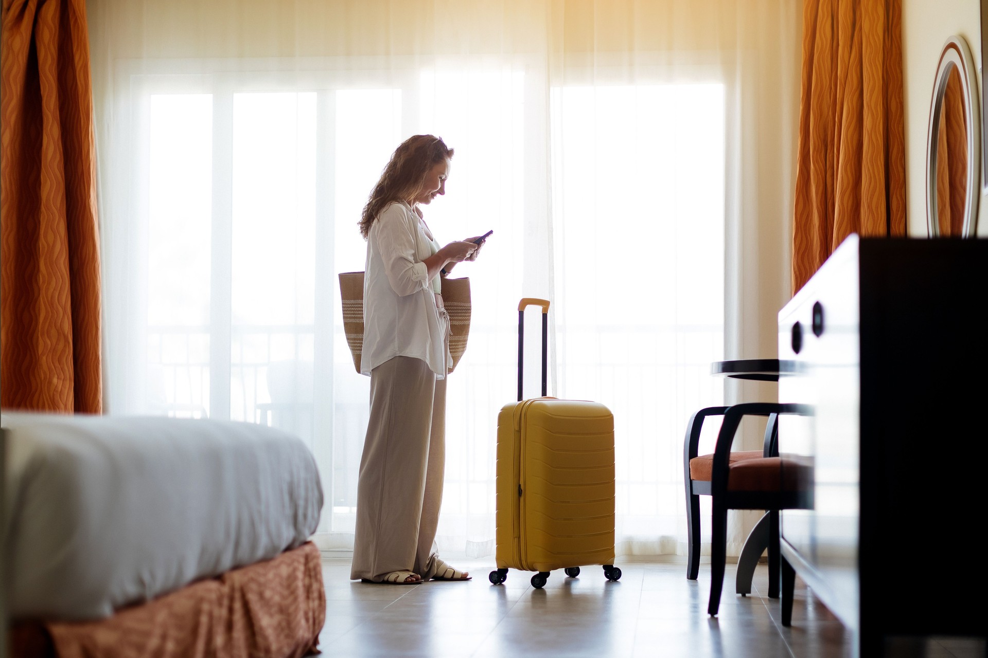 Woman standing in hotel room with yellow suitcase, looking at her phone